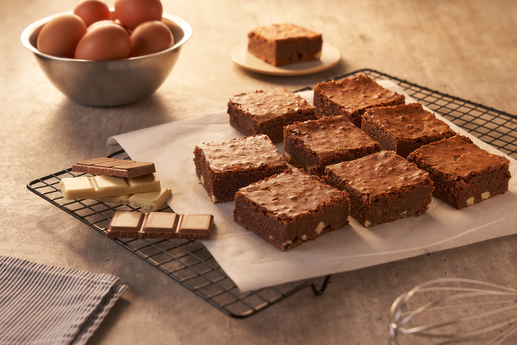 Nove brownies de chocolate com pedaços de nozes sobre papel manteiga em uma grade de resfriamento. Ao lado, barras de chocolate ao leite e branco, ovos em uma tigela e um batedor de arame, sugerindo preparo. Um brownie solitário está em um prato ao fundo