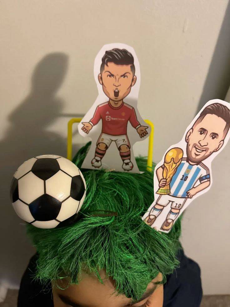 Decora&ccedil;&atilde;o de festa com um boneco de Cristiano Ronaldo em camisa vermelha e Messi em camisa azul e branca segurando a ta&ccedil;a, sobre uma peruca verde e uma bola de futebol