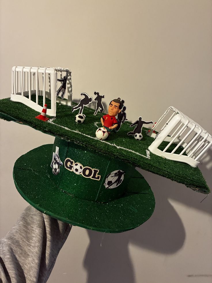 Chap&eacute;u verde brilhante com um campo de futebol em miniatura no topo, incluindo dois gols brancos, um cone laranja, um boneco de jogador de futebol com camisa vermelha e v&aacute;rios recortes de silhuetas de jogadores. A aba do chap&eacute;u tem a palavra GOOL com uma bola de futebol no lugar do O