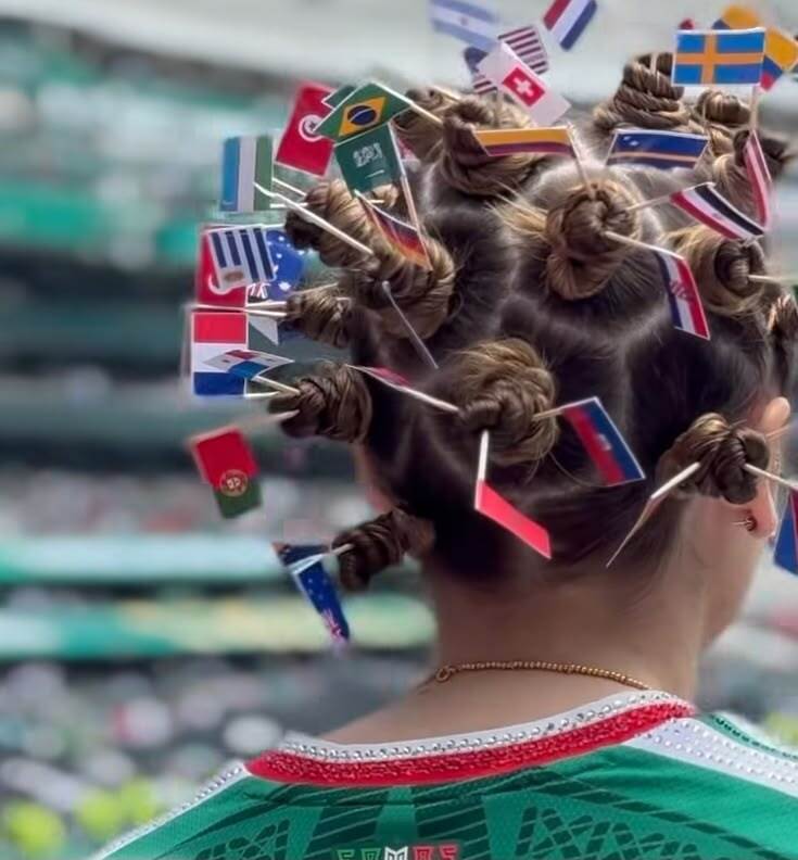 Pessoa de costas com cabelo preso em v&aacute;rios coques, cada um com uma pequena bandeira de pa&iacute;ses como Brasil, Ar&aacute;bia Saudita e Su&eacute;cia. Veste uma camisa verde com detalhes vermelhos e brancos na gola, e um colar dourado.