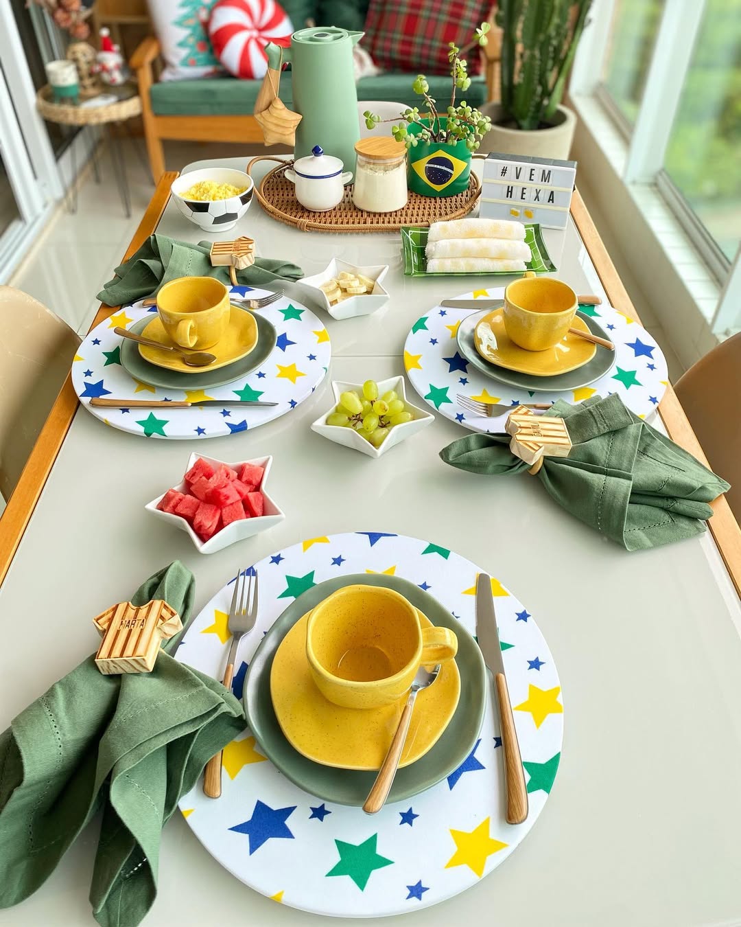 Mesa de caf&eacute; da manh&atilde; decorada com tema Brasil, incluindo jogos americanos de estrelas verdes, amarelas e azuis, x&iacute;caras amarelas, guardanapos verdes com an&eacute;is em formato de camisa de futebol, frutas em tigelas de estrela, tapiocas, e uma placa luminosa com a hashtag #VEMHEXA