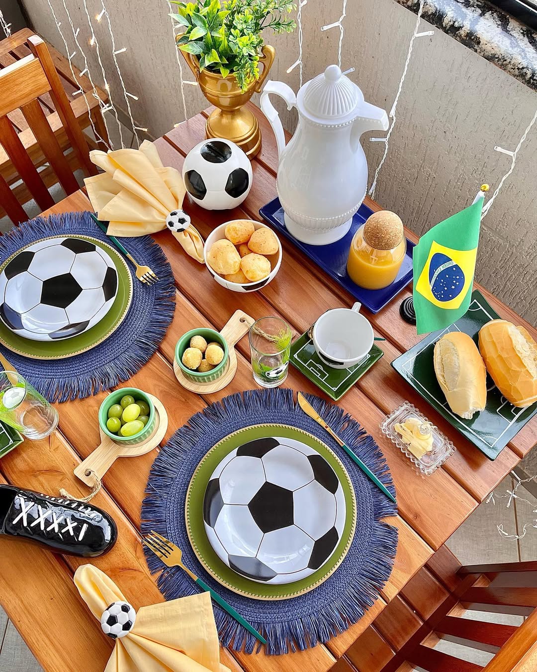 Mesa de madeira posta para caf&eacute; da manh&atilde; com tema de futebol, incluindo pratos de bola, guardanapos amarelos com an&eacute;is de bola, p&atilde;es de queijo, uvas, p&atilde;es franceses, manteiga, suco de laranja, garrafa t&eacute;rmica branca, ta&ccedil;a dourada com planta, chuteira preta decorativa e uma pequena bandeira do Brasil