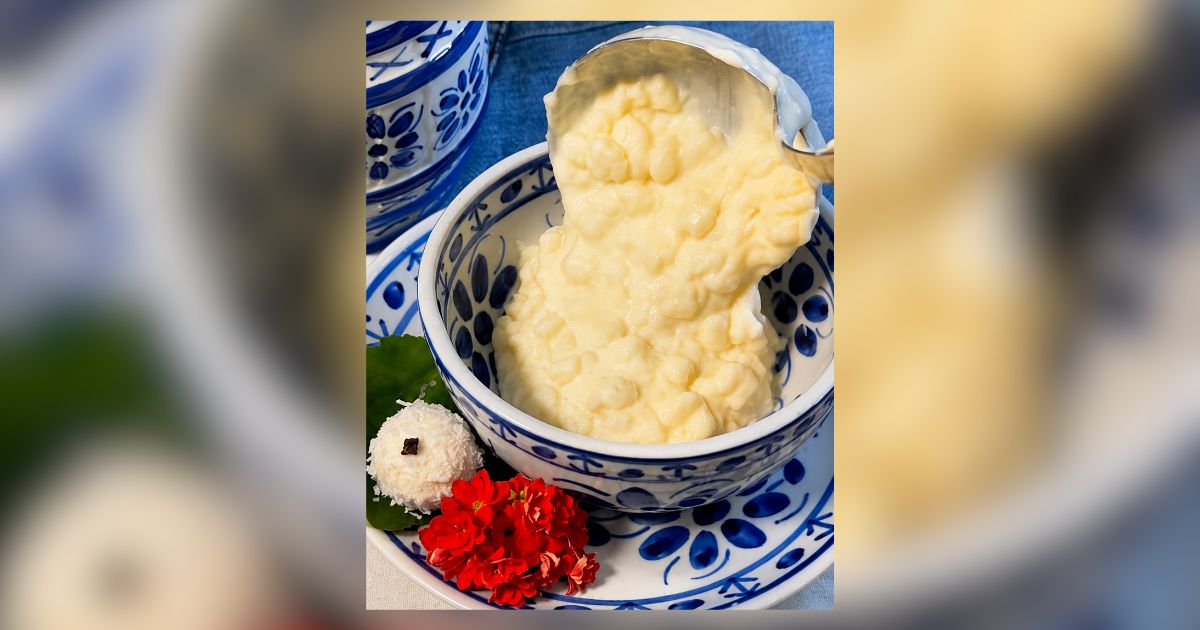 Uma concha de metal despeja um creme amarelo claro e espesso em uma tigela branca e azul. Ao lado, um doce de coco branco com um quadrado escuro no topo e flores vermelhas vibrantes sobre uma folha verde, tudo em um prato decorado com padrões azuis e brancos