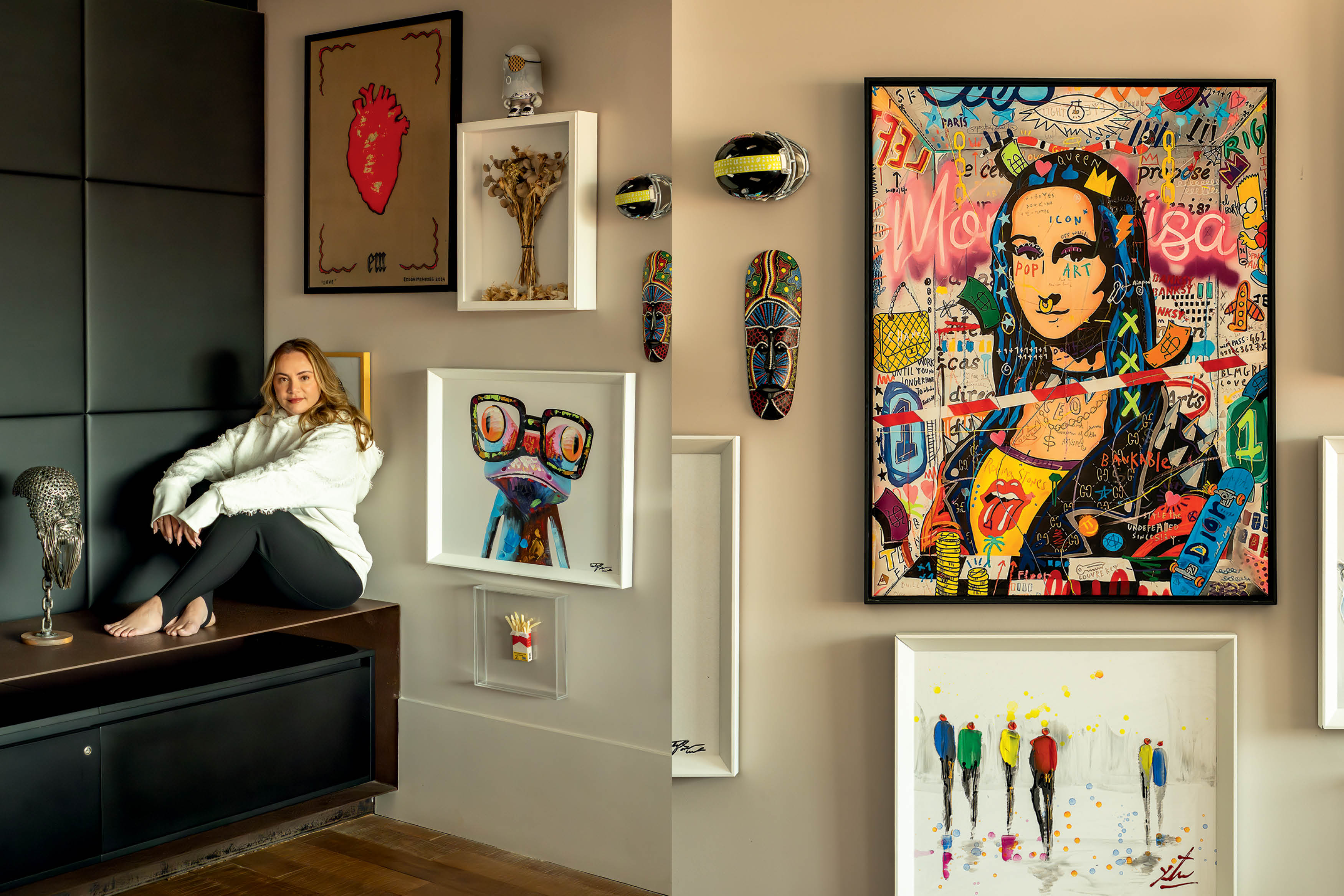 Mulher de cabelos loiros sentada descal&ccedil;a sobre um m&oacute;vel escuro, sorrindo, em um ambiente decorado com quadros de arte pop e m&aacute;scaras africanas