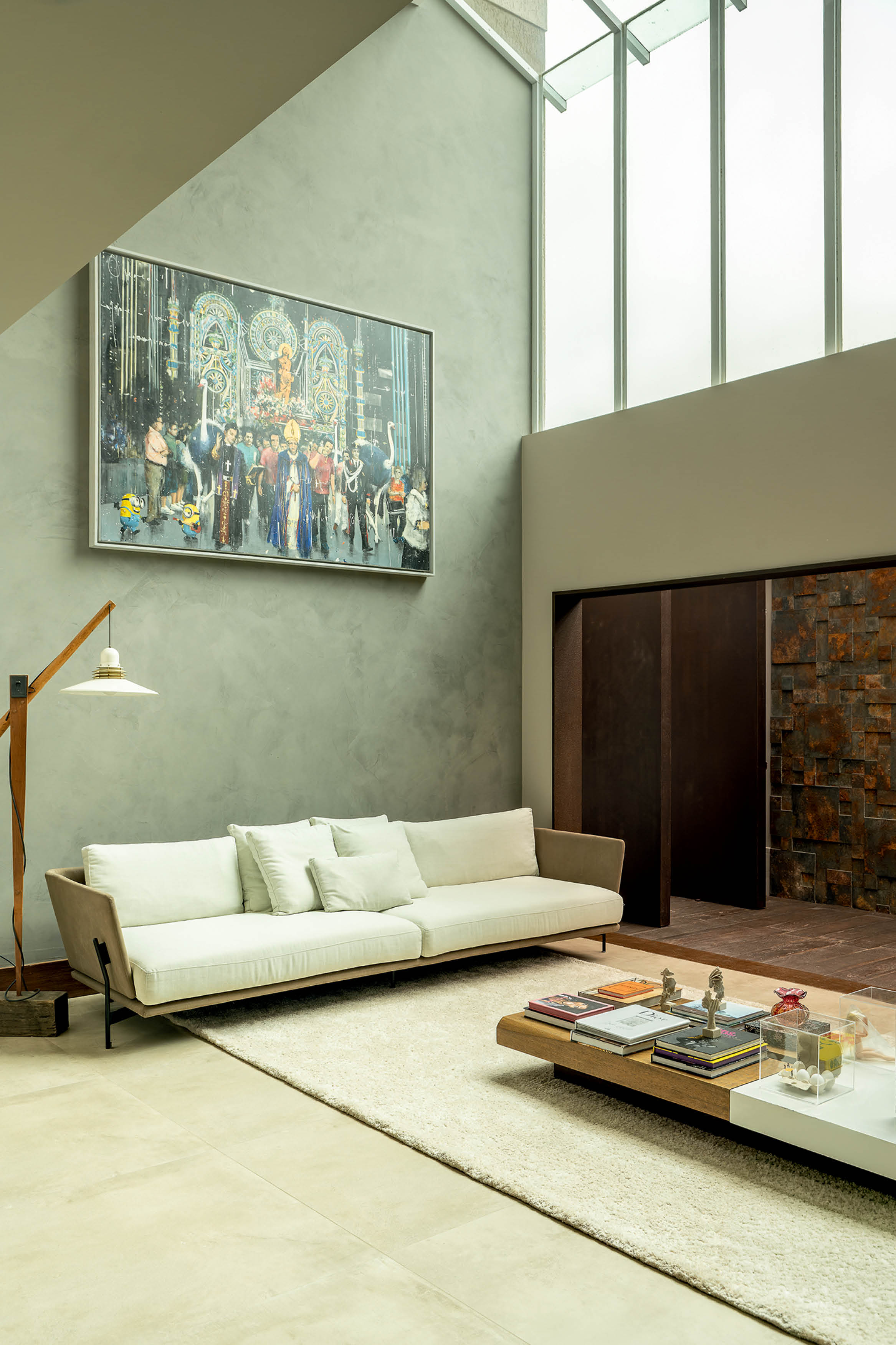 Sala de estar moderna com sof&aacute; bege e almofadas brancas sobre tapete felpudo claro. Na parede cinza, um quadro colorido de prociss&atilde;o com figuras religiosas e personagens de desenho. &Agrave; esquerda, lumin&aacute;ria de ch&atilde;o com base de madeira. &Agrave; direita, porta de correr escura e parede com revestimento de pedra r&uacute;stica. Janelas altas no teto trazem luz natural.