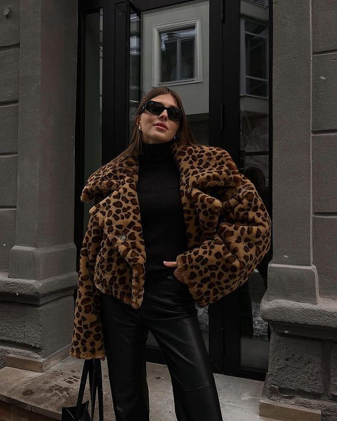 Mulher jovem, de cabelos castanhos longos, usando &oacute;culos de sol retangulares, blusa preta de gola alta, casaco de pele sint&eacute;tica com estampa de leopardo e cal&ccedil;a preta de couro, posando em frente a uma porta escura e parede cinza de um edif&iacute;cio
