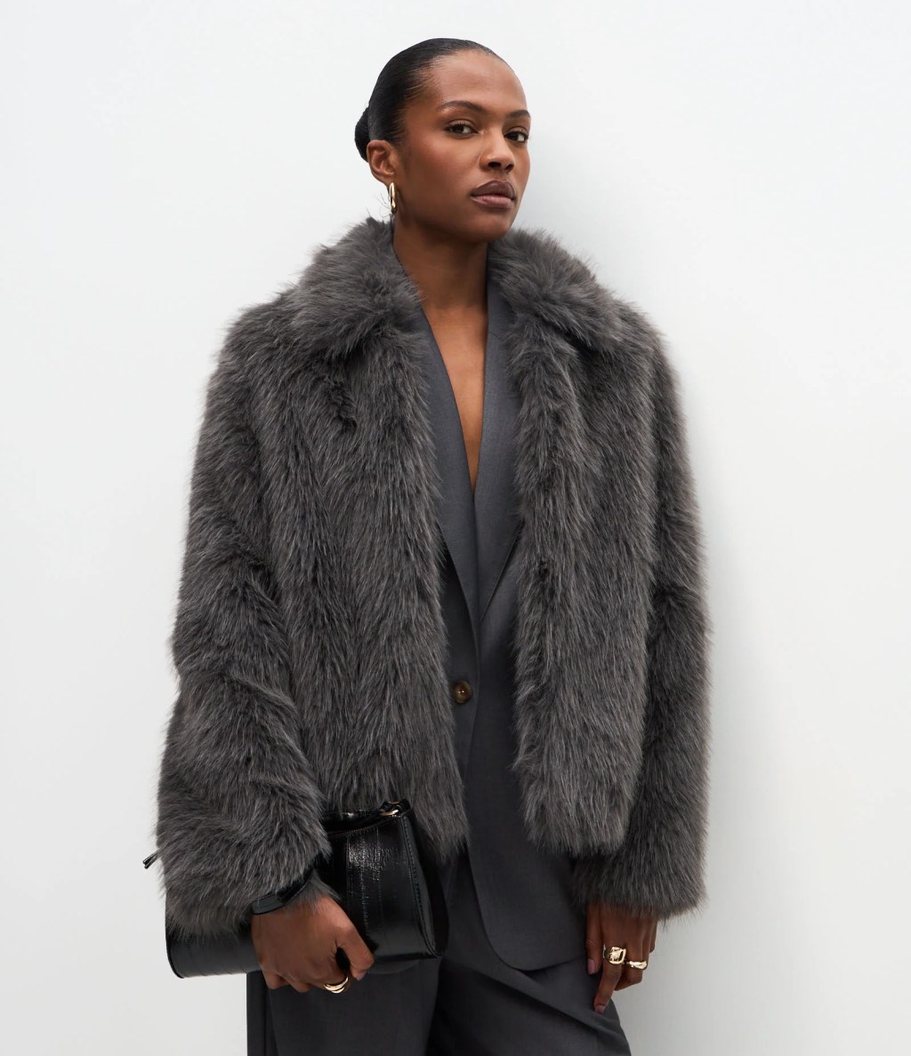 Mulher negra com coque, brincos dourados e express&atilde;o s&eacute;ria, vestindo casaco cinza de pelos sobre blazer e cal&ccedil;a cinzas, segurando uma bolsa preta de couro texturizado. Ela usa an&eacute;is dourados e est&aacute; em frente a uma parede branca