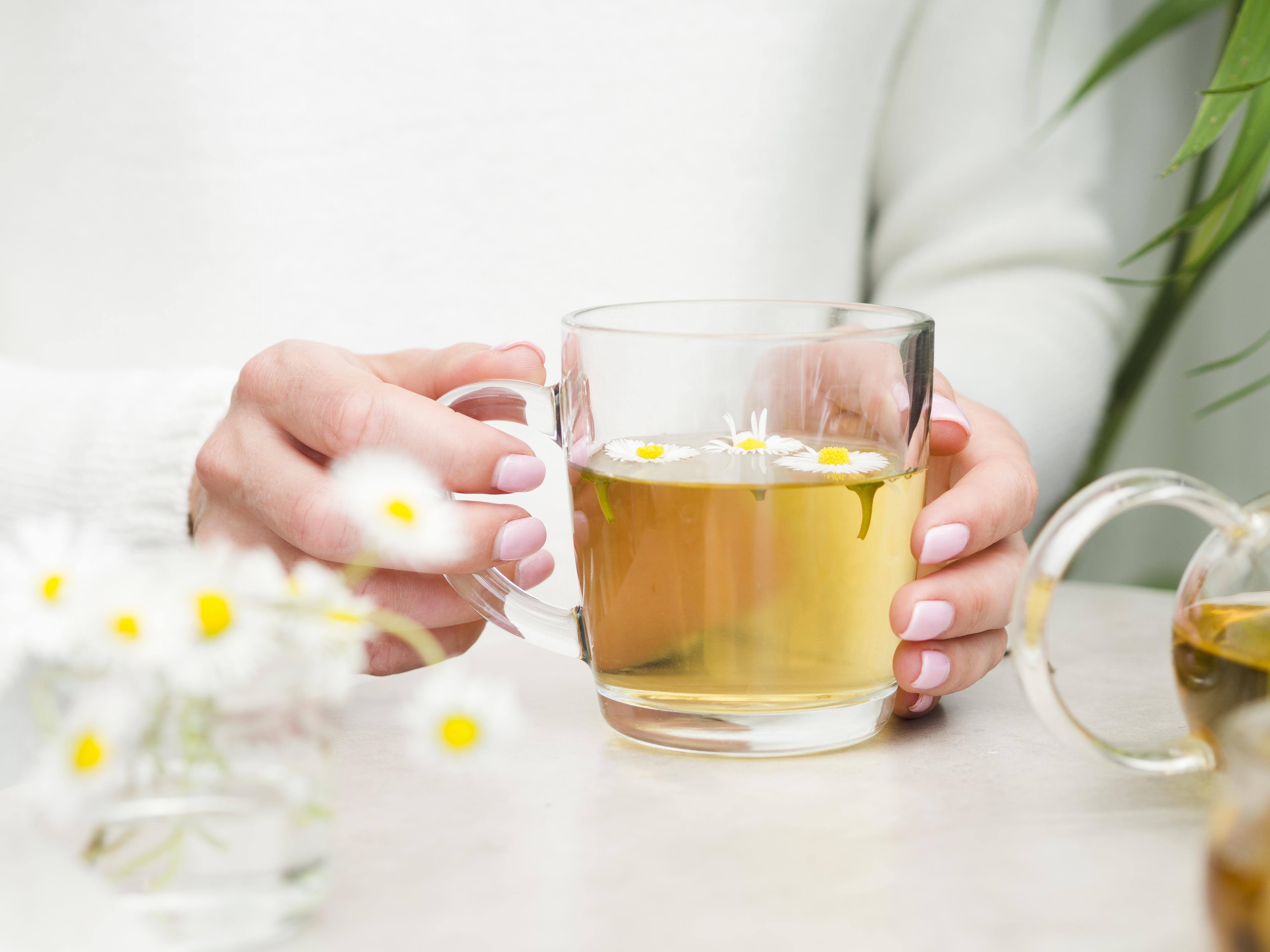 M&atilde;os femininas com unhas rosadas seguram uma x&iacute;cara de ch&aacute; de camomila com flores flutuando, ao lado de um buqu&ecirc; de margaridas e um bule de vidro