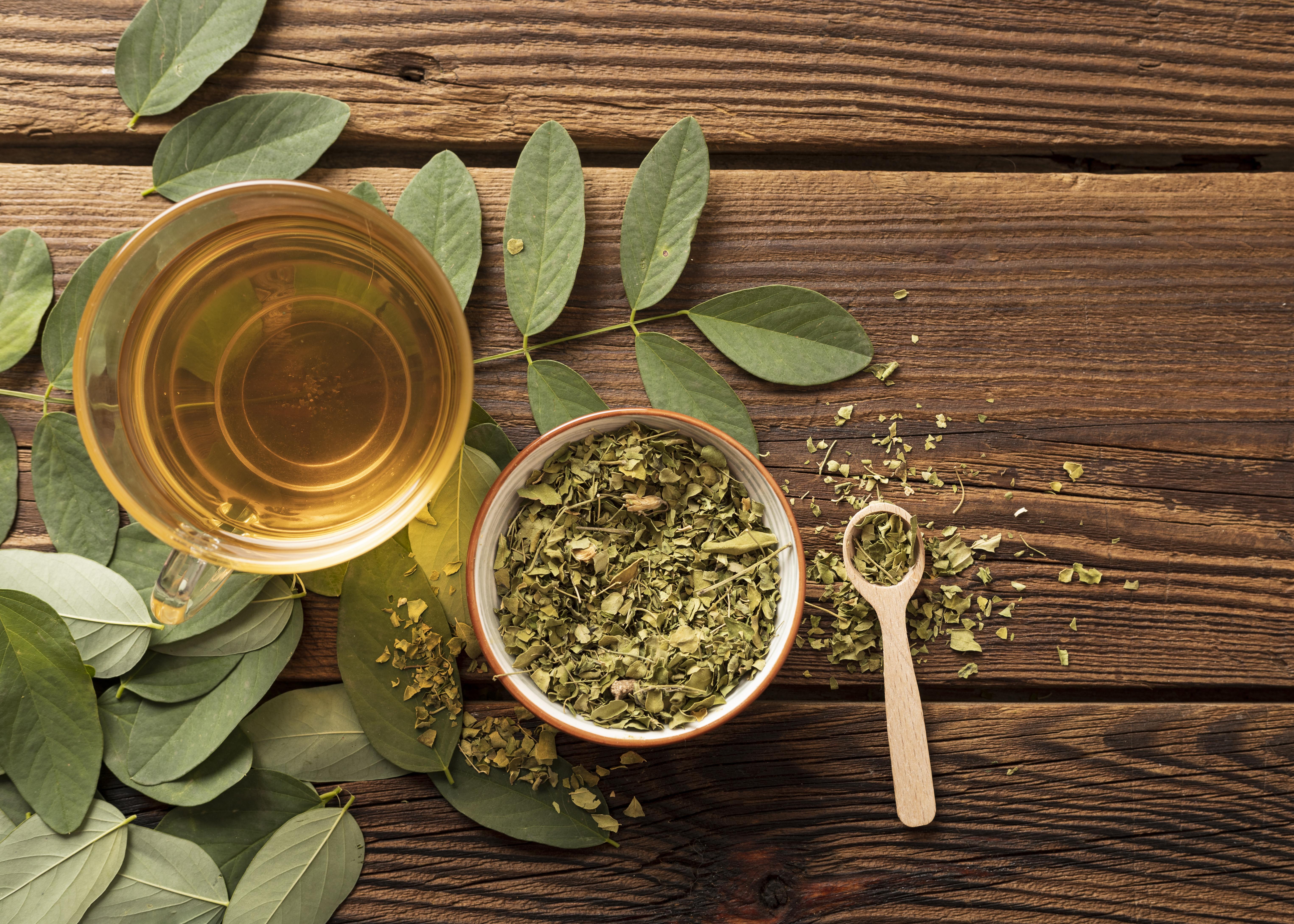 X&iacute;cara de ch&aacute; dourado, folhas verdes frescas e secas em uma tigela e colher de pau sobre mesa r&uacute;stica de madeira