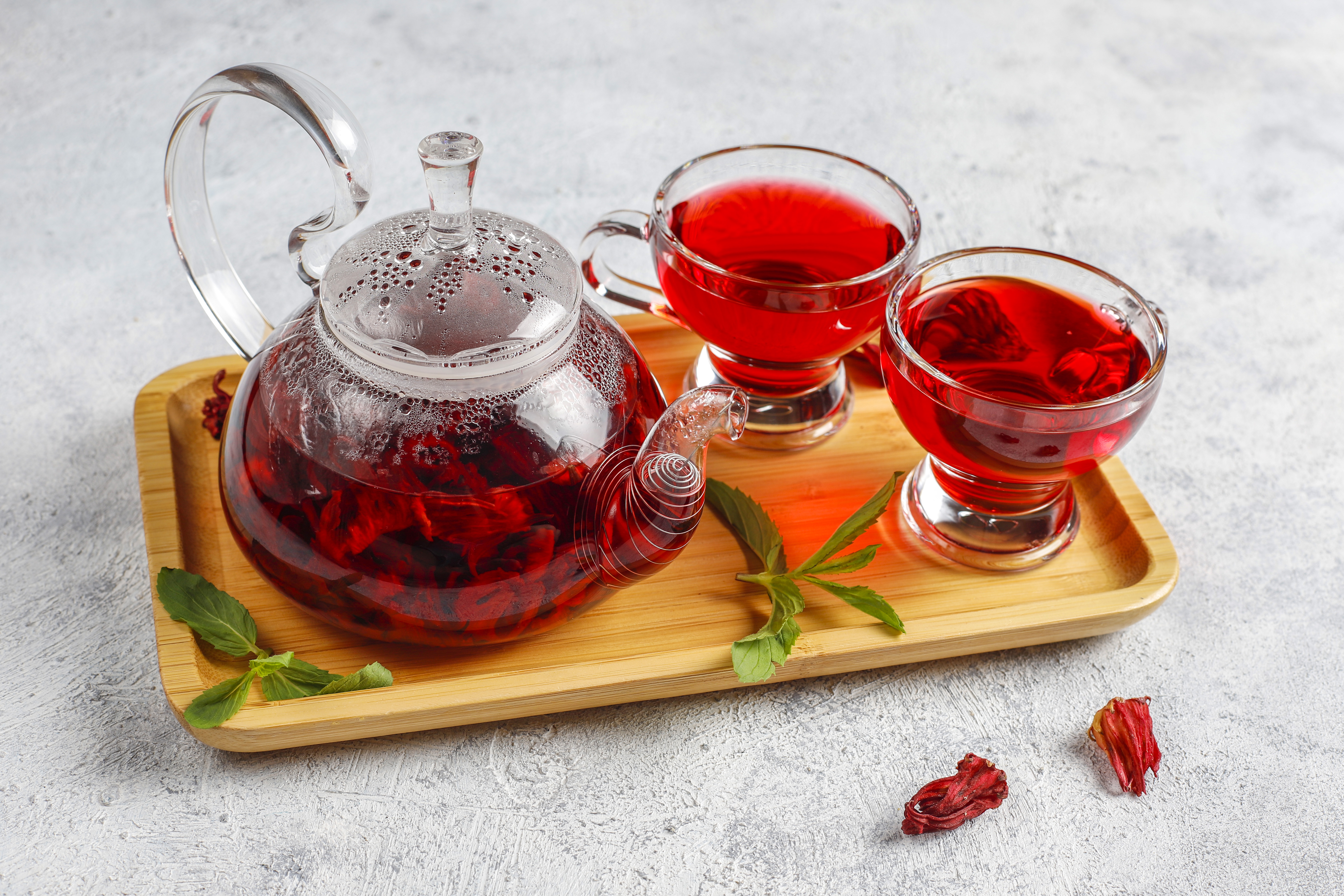 Bule de vidro transparente com ch&aacute; de hibisco vermelho intenso e duas x&iacute;caras de ch&aacute;, sobre uma bandeja de bambu. Folhas de hortel&atilde; e flores secas de hibisco est&atilde;o espalhadas na bandeja e na superf&iacute;cie cinza