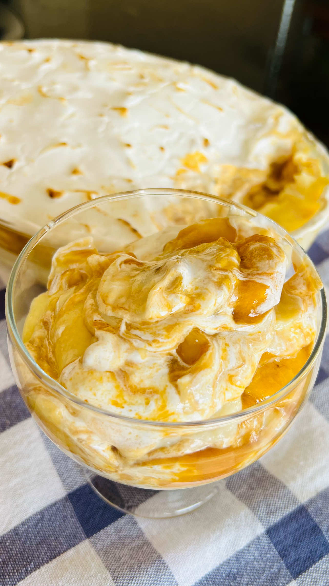 Uma ta&ccedil;a de vidro com doce de p&ecirc;ssego em calda e creme branco, com um tabuleiro do mesmo doce ao fundo, sobre um pano de prato xadrez azul e branco