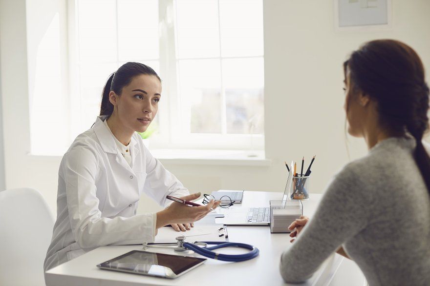M&eacute;dica jovem de jaleco branco, cabelo preso, conversando com paciente de costas, em consult&oacute;rio claro com janela, mesa com tablet, estetosc&oacute;pio e laptop