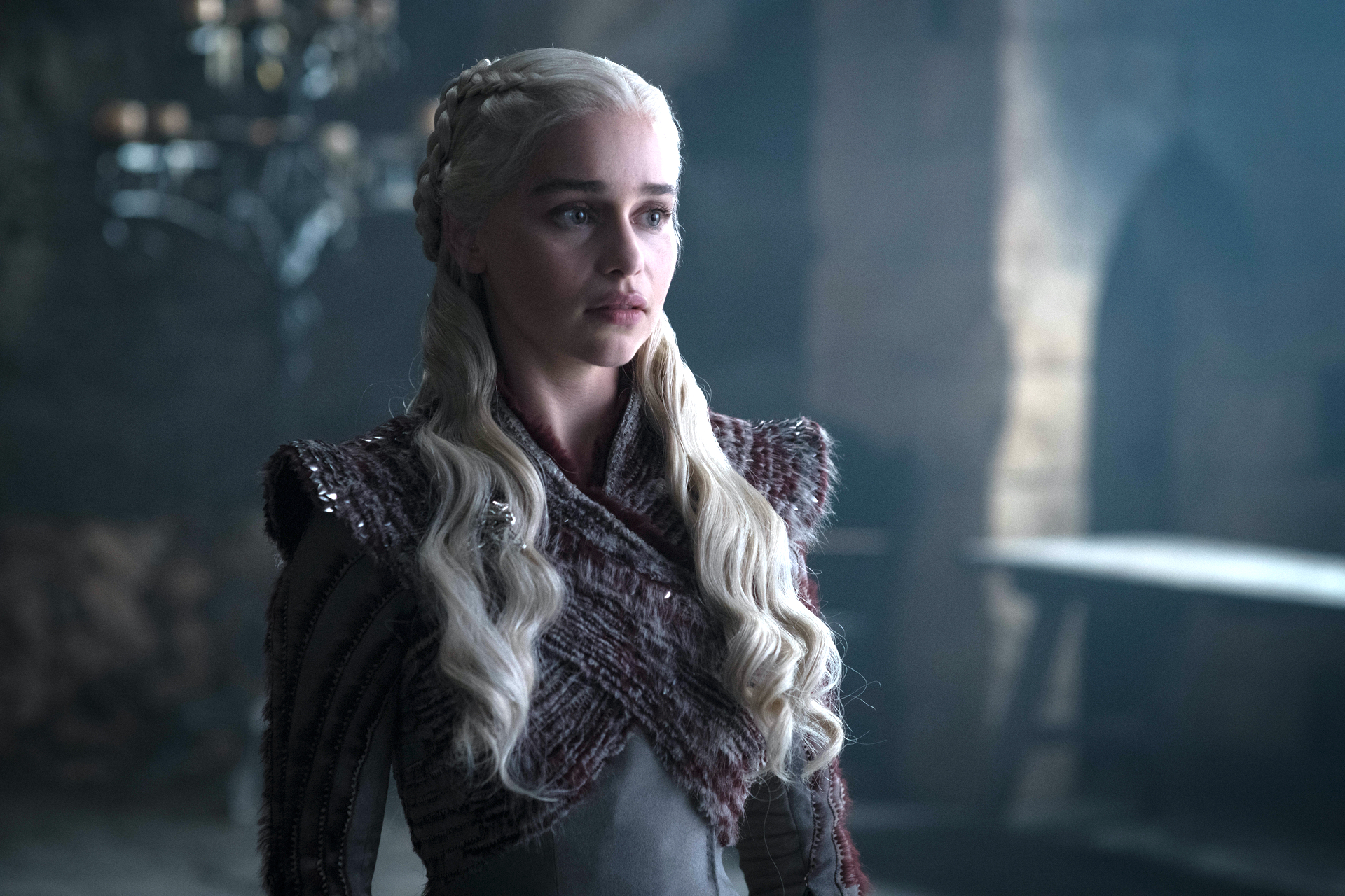 Mulher de cabelos loiros platinados, longos e ondulados, com tran&ccedil;as laterais, veste um casaco texturizado em tons de vinho e cinza, sobre uma roupa escura. Ela tem uma express&atilde;o s&eacute;ria, olhando para o lado esquerdo da imagem, em um ambiente escuro com ilumina&ccedil;&atilde;o difusa e um lustre ao fundo