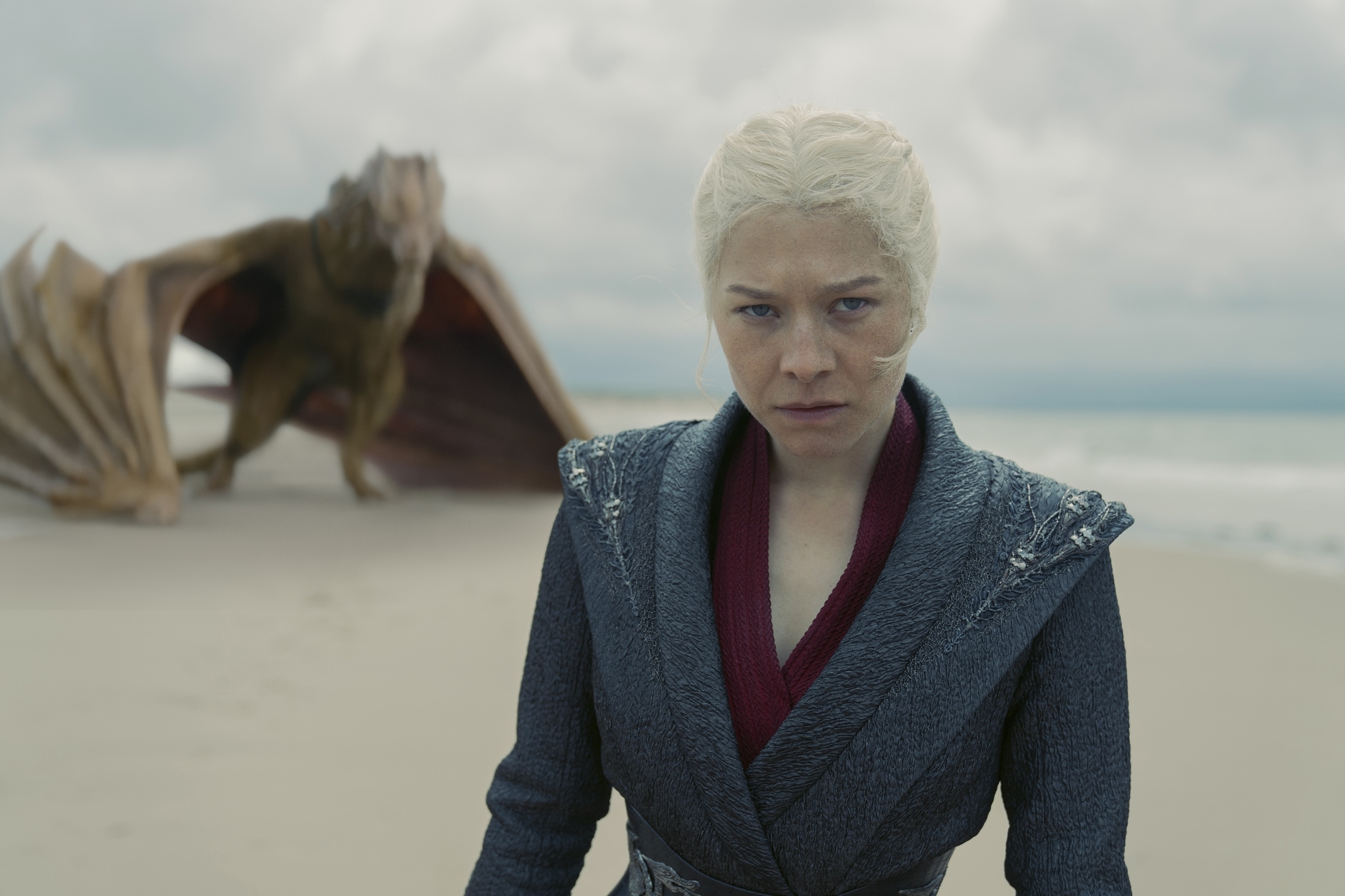 Uma mulher de cabelos loiros platinados e express&atilde;o s&eacute;ria, vestindo um casaco cinza escuro sobre uma t&uacute;nica vinho, olha diretamente para frente. Ao fundo, um drag&atilde;o marrom com asas abertas est&aacute; em uma praia de areia clara, com o mar e o c&eacute;u nublado ao longe