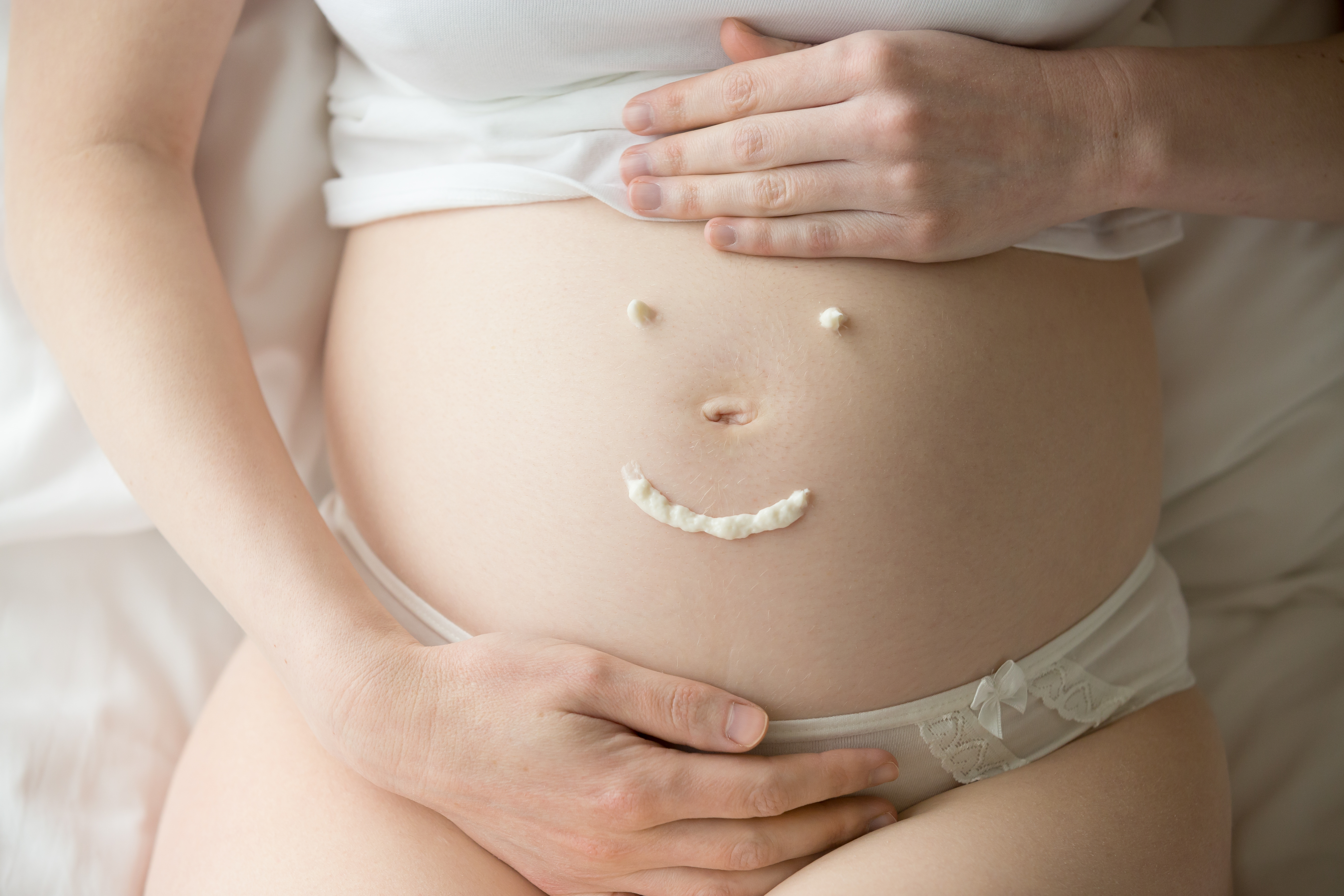 Barriga de uma mulher grávida com um rosto sorridente desenhado com creme, usando calcinha branca de renda e as mãos sobre a barriga