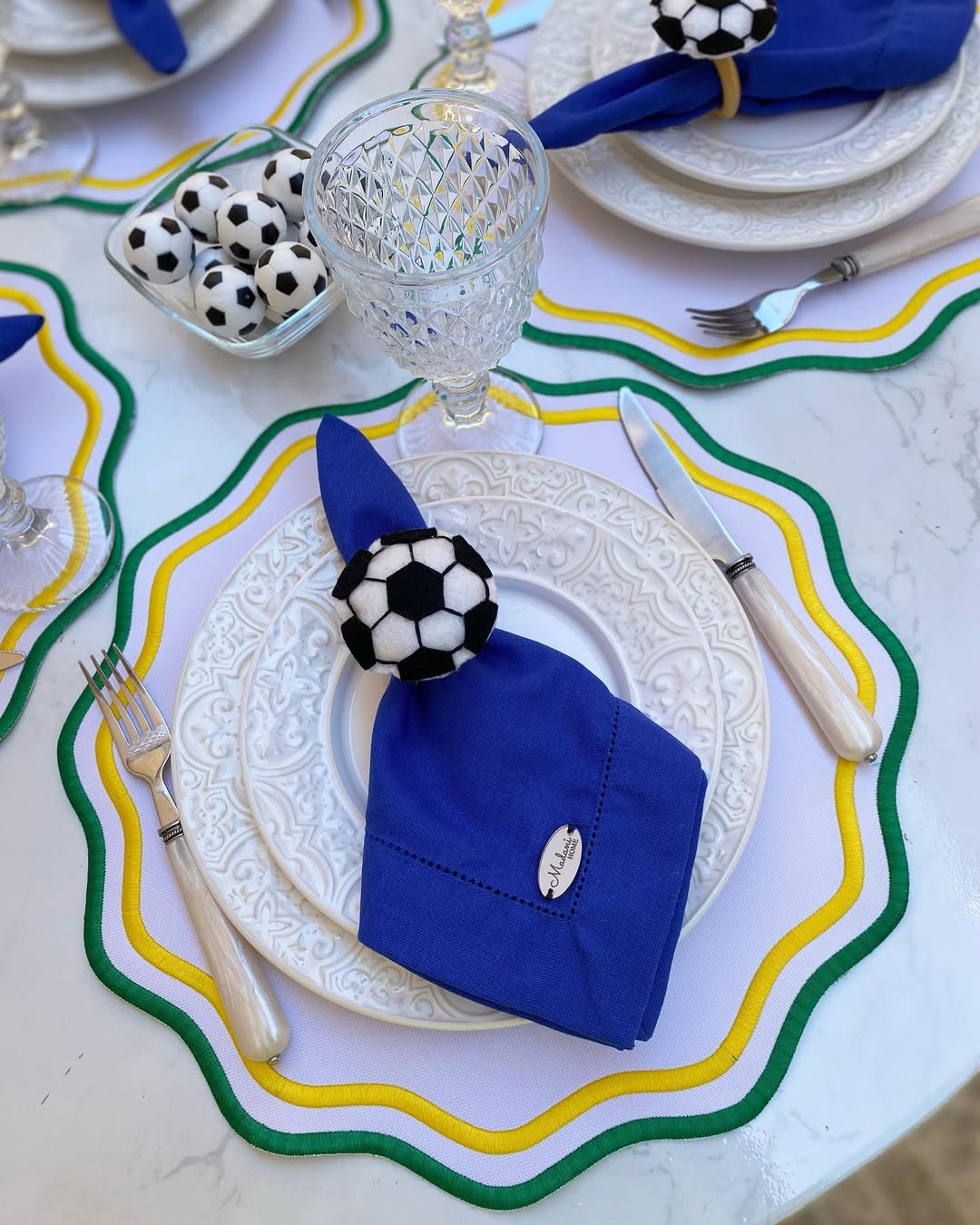 Mesa posta com pratos brancos decorados, guardanapos azuis presos por an&eacute;is de bola de futebol, talheres e copos de cristal. Jogos americanos brancos com bordas onduladas em verde, amarelo e branco. Uma tigela com pequenas bolas de futebol completa a decora&ccedil;&atilde;o tem&aacute;tica