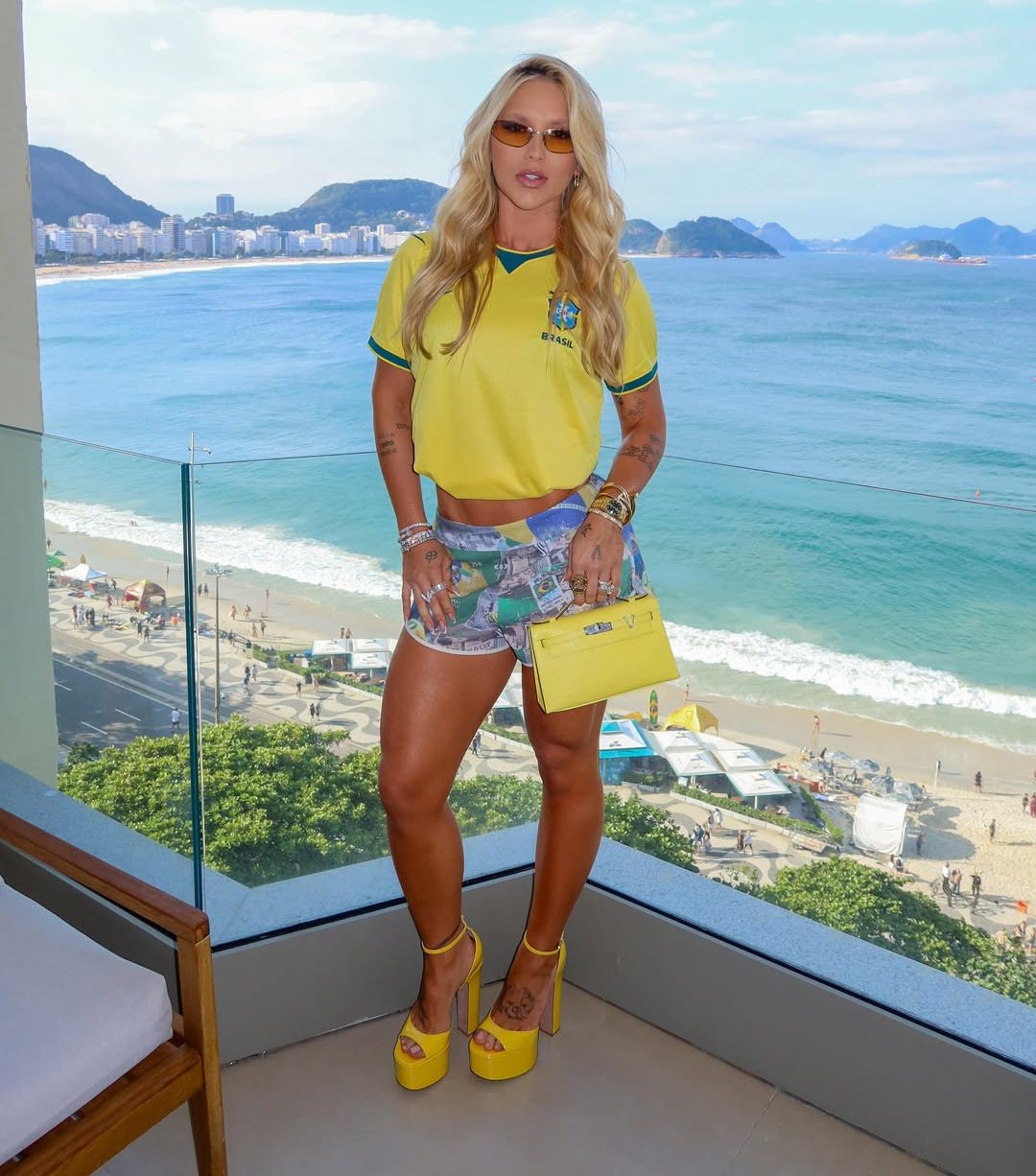 Mulher loira, de &oacute;culos escuros e camisa amarela da sele&ccedil;&atilde;o brasileira, posa em varanda com vista para a praia e o mar do Rio de Janeiro