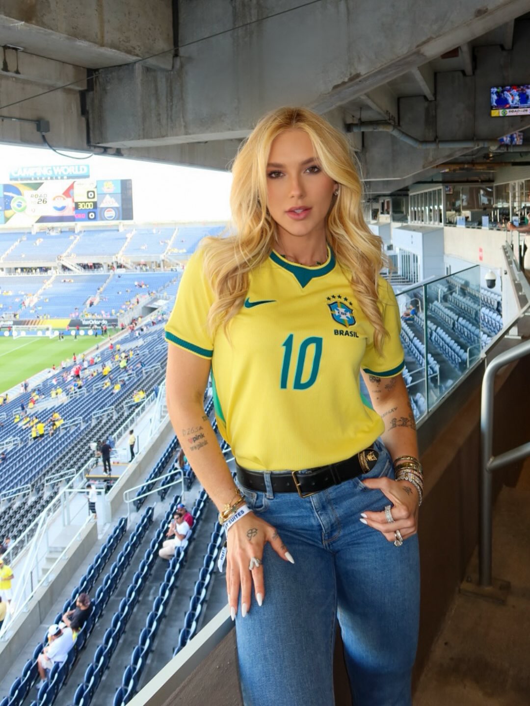Mulher loira, de cabelos ondulados, usando camisa amarela da sele&ccedil;&atilde;o brasileira com o n&uacute;mero 10 e cal&ccedil;a jeans, posa em um est&aacute;dio de futebol com arquibancadas azuis ao fundo