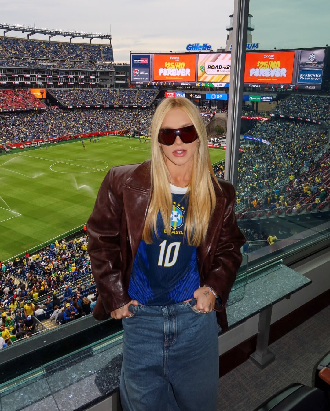 Mulher loira de &oacute;culos escuros e jaqueta marrom sobre camisa da sele&ccedil;&atilde;o brasileira n&uacute;mero 10, posando em um camarote de est&aacute;dio de futebol lotado