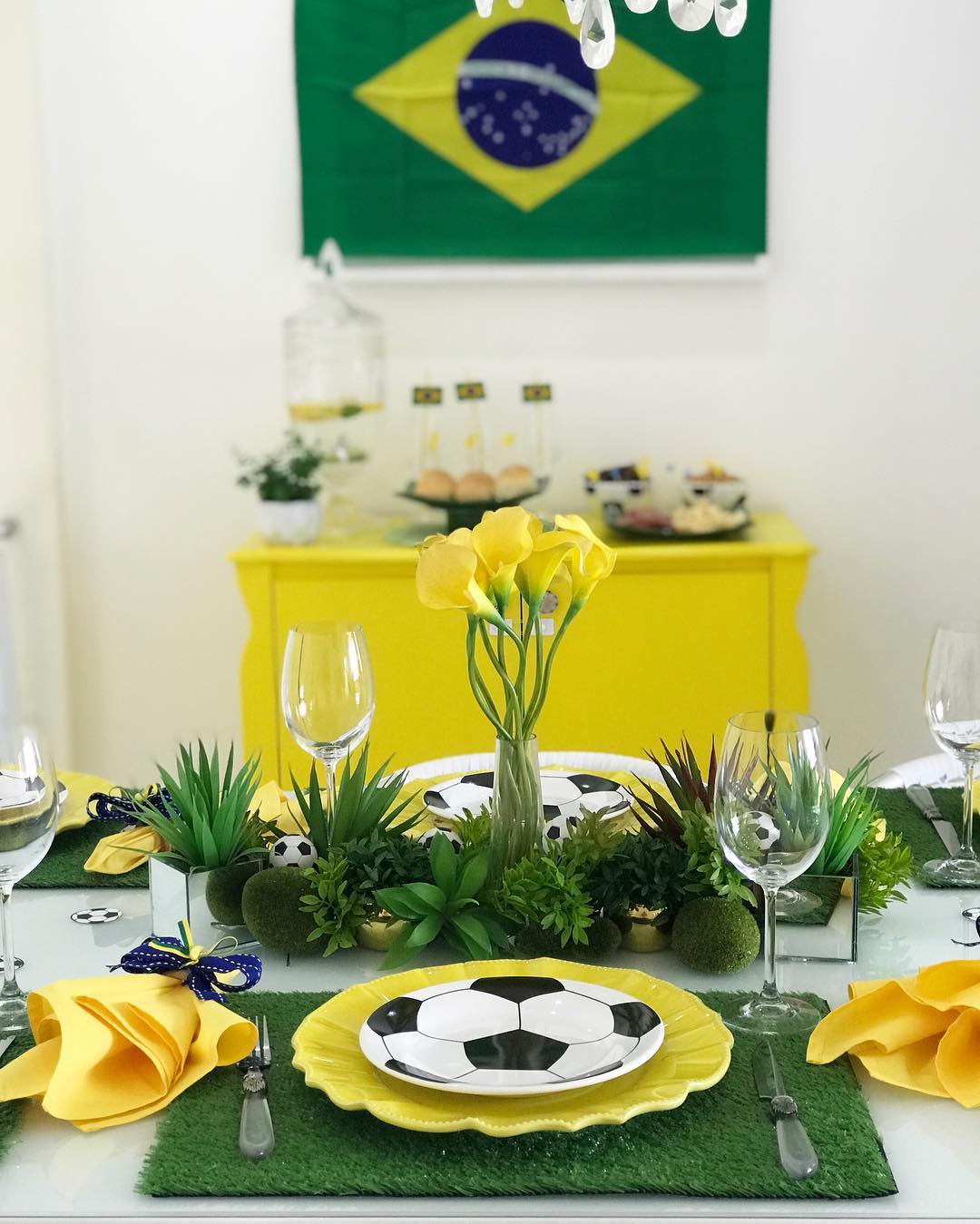 Mesa decorada para festa tem&aacute;tica de futebol, com toalhas de mesa imitando gramado, pratos amarelos e pratos menores com estampa de bola de futebol. H&aacute; um arranjo de flores amarelas e folhagens verdes no centro, ta&ccedil;as de vinho e guardanapos amarelos presos com la&ccedil;os azuis e amarelos. Ao fundo, uma c&ocirc;moda amarela com doces e uma bandeira do Brasil pendurada na parede