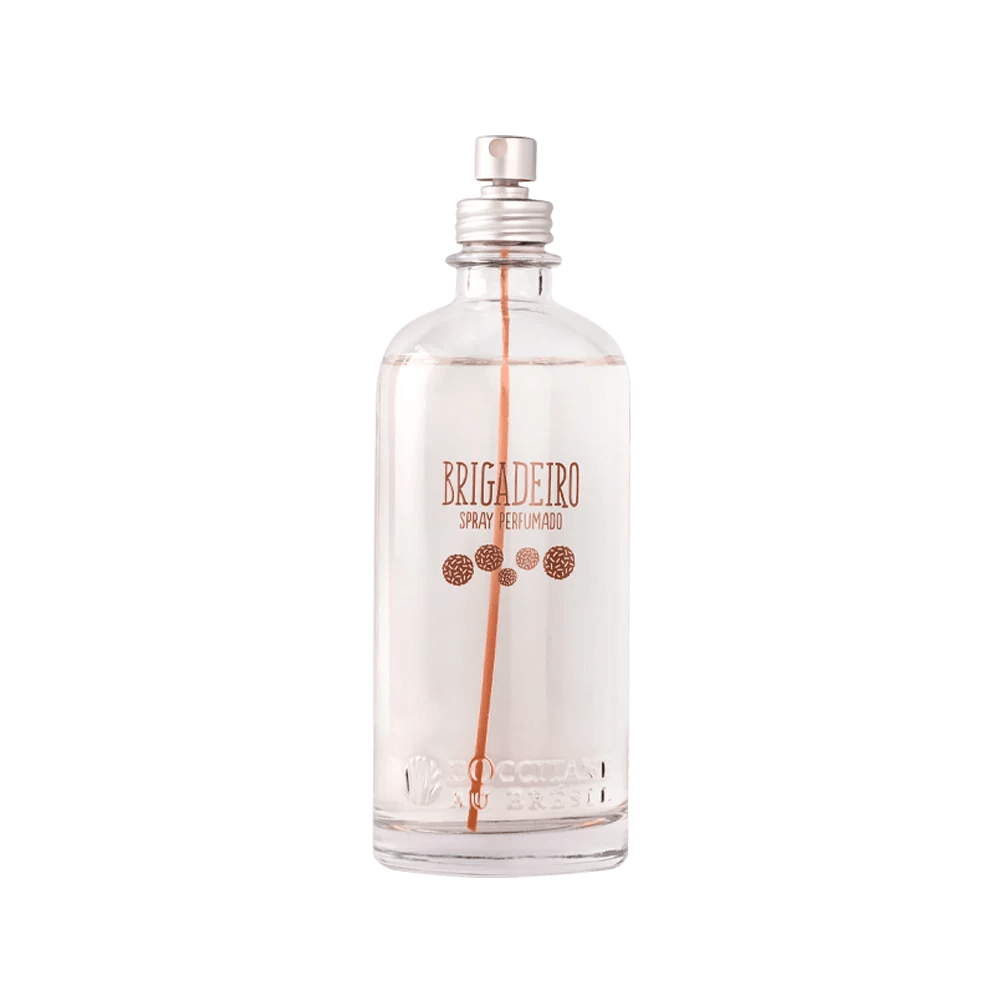 Frasco transparente de spray perfumado Brigadeiro da LOccitane Au Br&eacute;sil, com l&iacute;quido claro e tubo interno laranja. O r&oacute;tulo tem letras em tom cobre e desenhos de granulados de brigadeiro