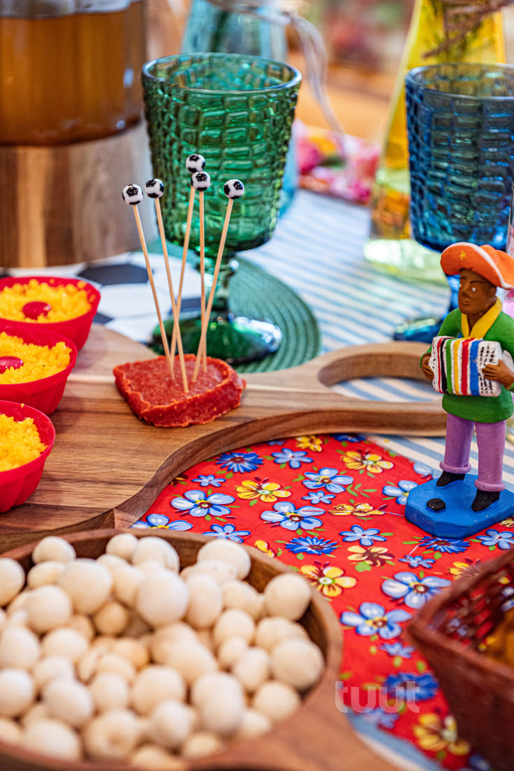 Mesa de festa junina com petiscos e decora&ccedil;&atilde;o colorida. Em primeiro plano, uma tigela de madeira com amendoins brancos. Ao fundo, uma t&aacute;bua de madeira com tr&ecirc;s potes vermelhos de cuscuz e um peda&ccedil;o de carne vermelha com espetos de bolinhas de futebol. &Agrave; direita, um boneco de sanfoneiro e uma toalha de mesa vermelha com flores azuis e amarelas. Copos verdes e azuis completam a cena festiva