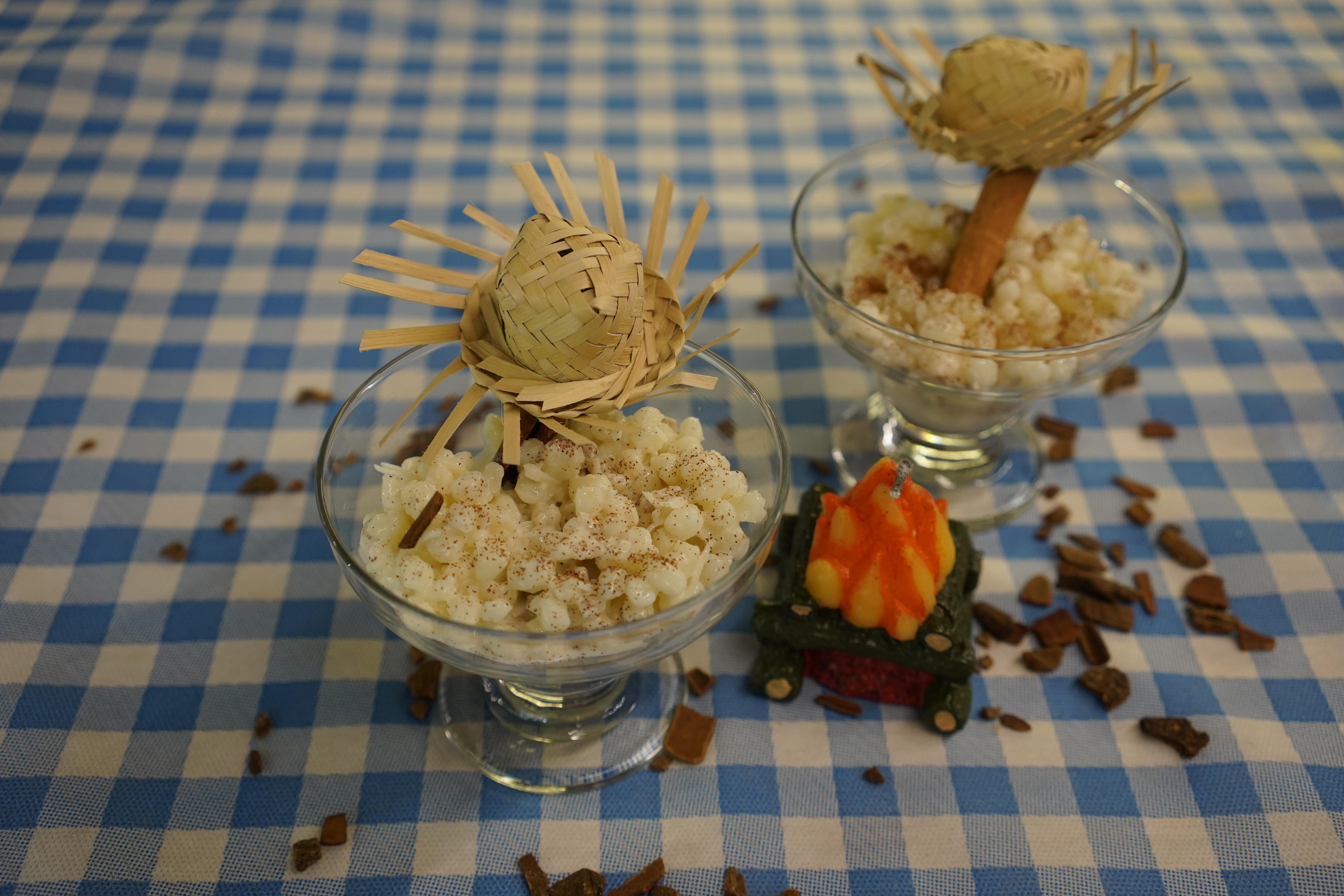 Duas ta&ccedil;as de curau de milho branco com canela em p&oacute;, decoradas com chap&eacute;us de palha em miniatura, sobre uma toalha xadrez azul e branca, com uma vela em formato de fogueira e peda&ccedil;os de canela em pau espalhados