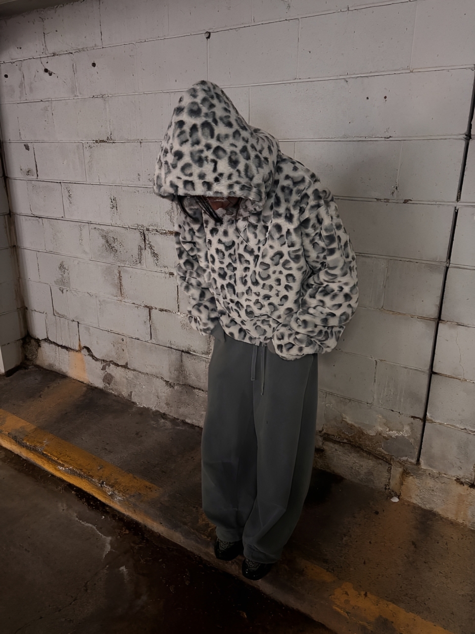 Pessoa com moletom felpudo de estampa de leopardo cinza e branco, capuz cobrindo o rosto, cal&ccedil;a larga cinza-escura e t&ecirc;nis pretos, em um estacionamento com parede de blocos de concreto e ch&atilde;o &uacute;mido com faixa amarela