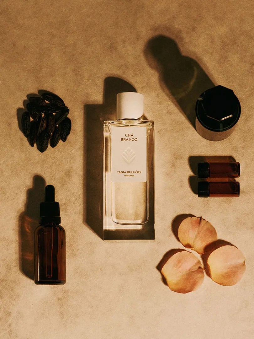 Frasco de perfume Ch&aacute; Branco Tania Bulh&otilde;es com tampa branca, cercado por gr&atilde;os escuros, um frasco conta-gotas, tr&ecirc;s p&eacute;talas de rosa e outros recipientes sobre uma superf&iacute;cie bege, com sombras alongadas