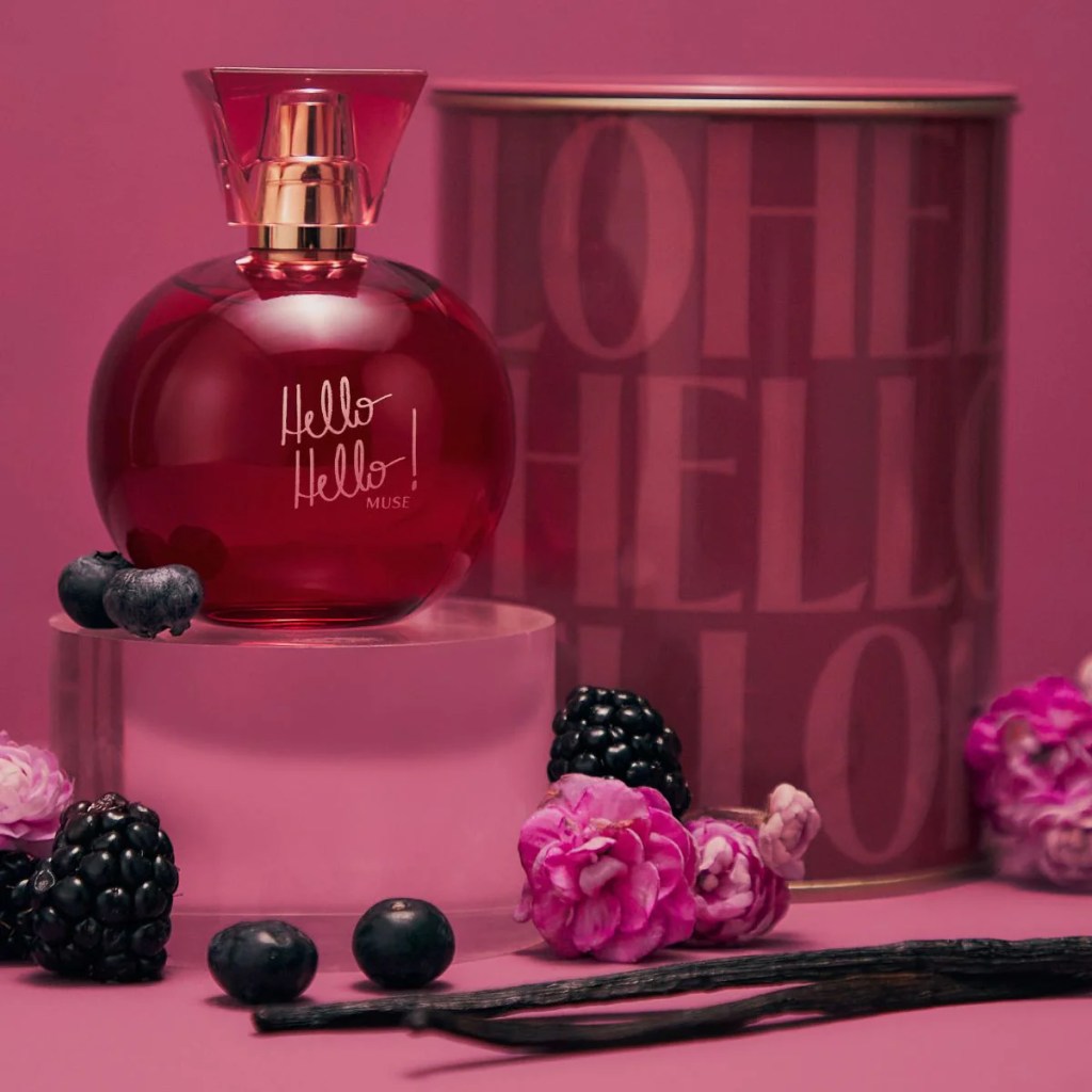 Frasco de perfume esf&eacute;rico vermelho com tampa dourada e transparente, escrito Hello Hello! MUSE, sobre um pedestal transl&uacute;cido. Ao redor, mirtilos, amoras, flores cor-de-rosa e favas de baunilha escuras, em fundo rosa com lata decorada