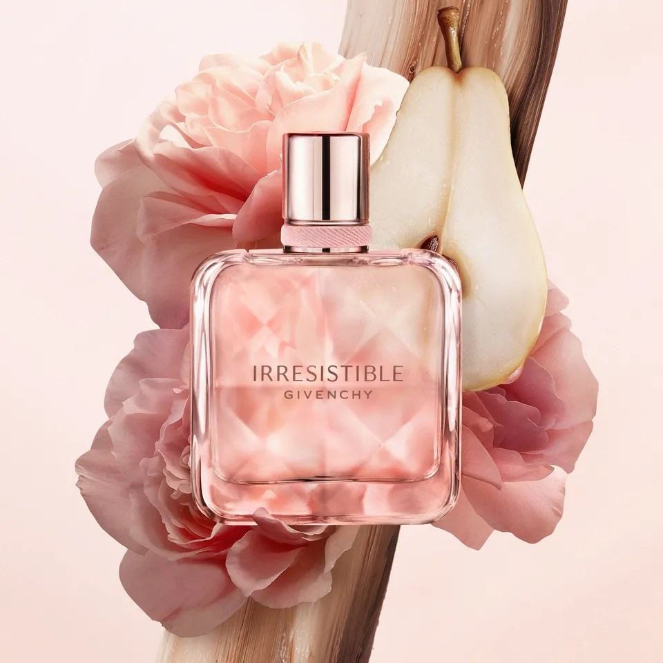 Frasco de perfume Irresistible Givenchy rosa claro com tampa met&aacute;lica, rodeado por p&eacute;talas de flores rosadas, uma pera cortada ao meio e um elemento marrom texturizado, em fundo rosa p&aacute;lido