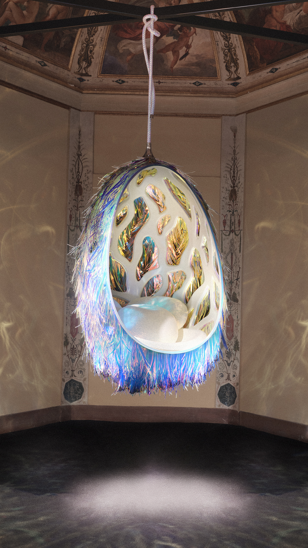 Cadeira suspensa em formato de ovo, com exterior de franjas iridescentes azuis e roxas, e interior branco com recortes de folhas coloridas. Almofadas brancas no assento. Pendurada por uma corda branca em um teto com afrescos cl&aacute;ssicos de anjos e figuras mitol&oacute;gicas. O ch&atilde;o &eacute; escuro, com reflexos de luz.