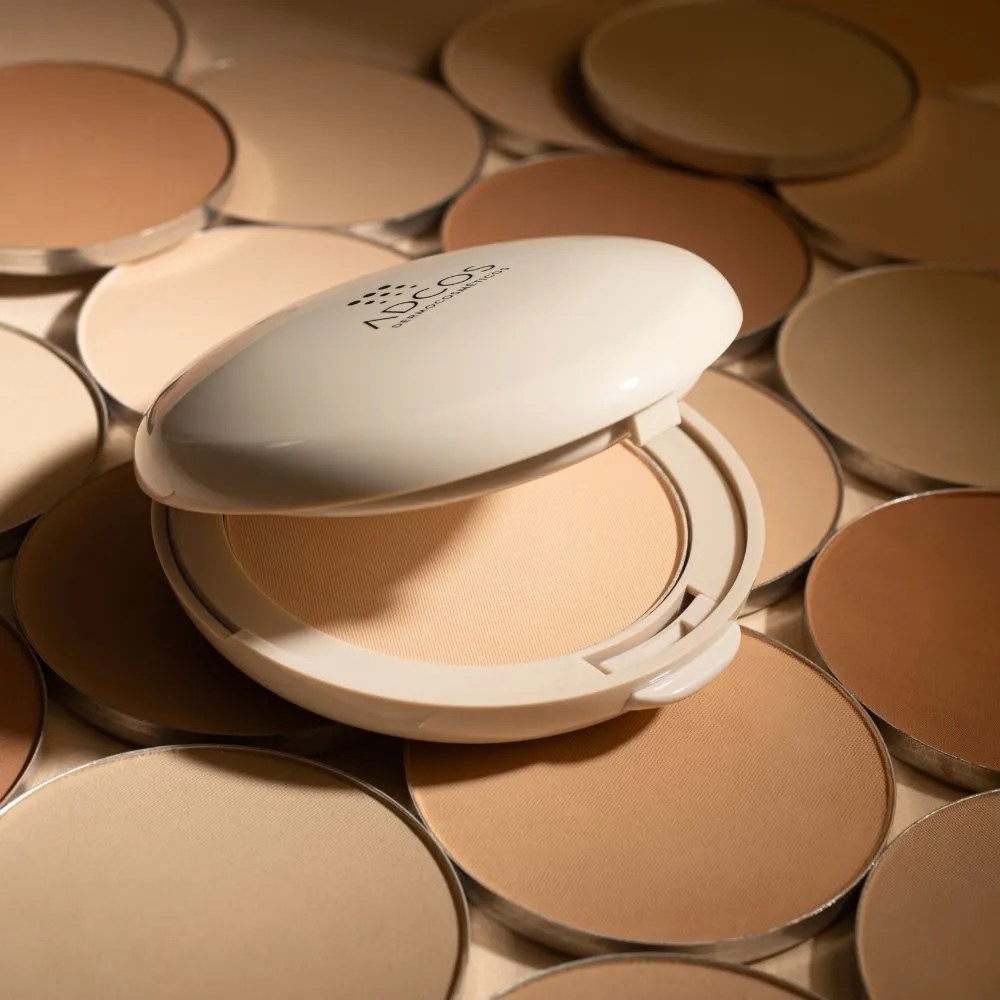 Um p&oacute; compacto bege claro aberto, com a tampa branca exibindo o logo NEOS DERMOCOSM&Eacute;TICOS, cercado por diversos refis de p&oacute; compacto em tons de pele variados, do claro ao escuro