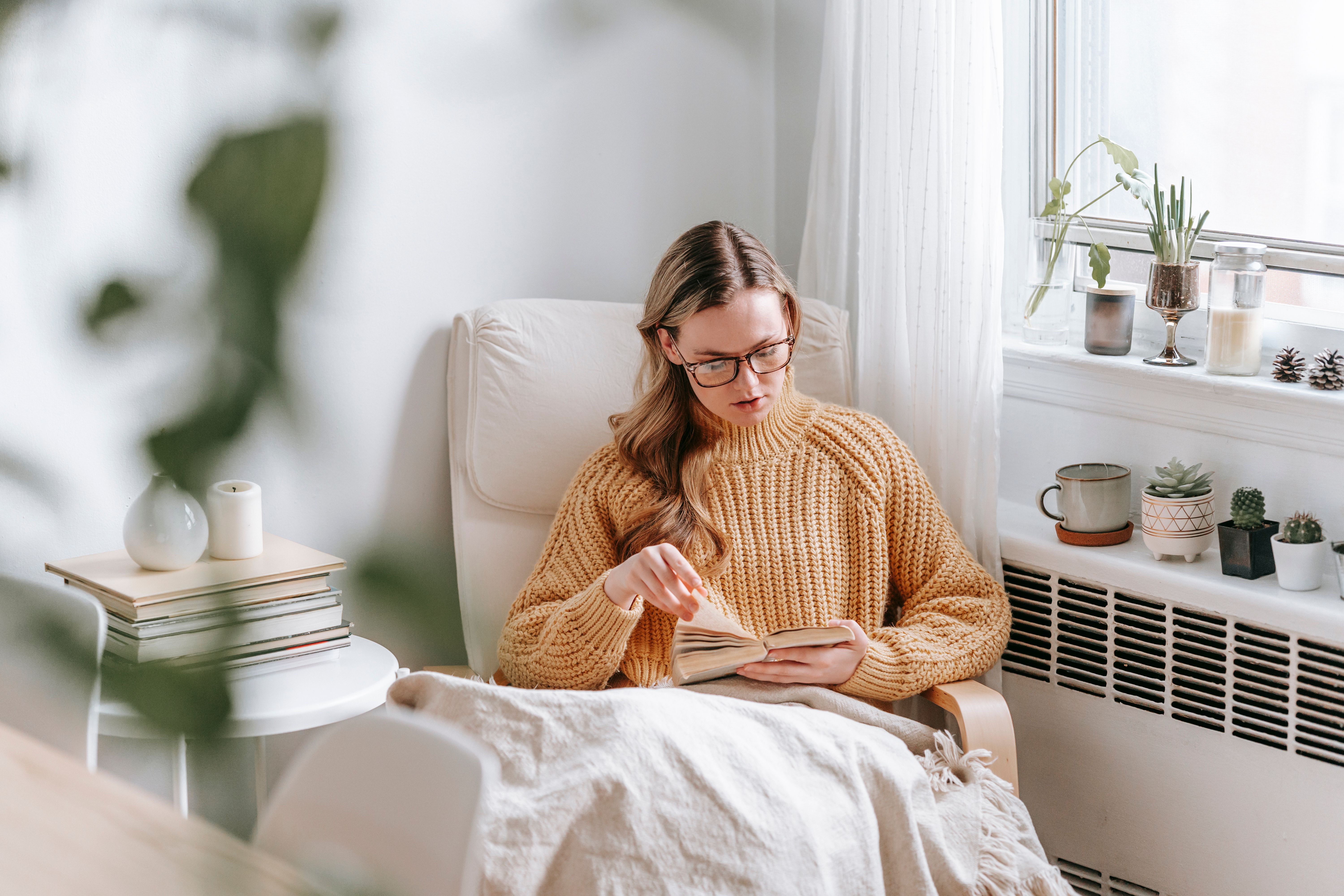 Mulher de óculos e suéter mostarda sentada em poltrona clara, lendo um livro. Ela está coberta por um cobertor bege, em um ambiente iluminado com plantas e velas na janela