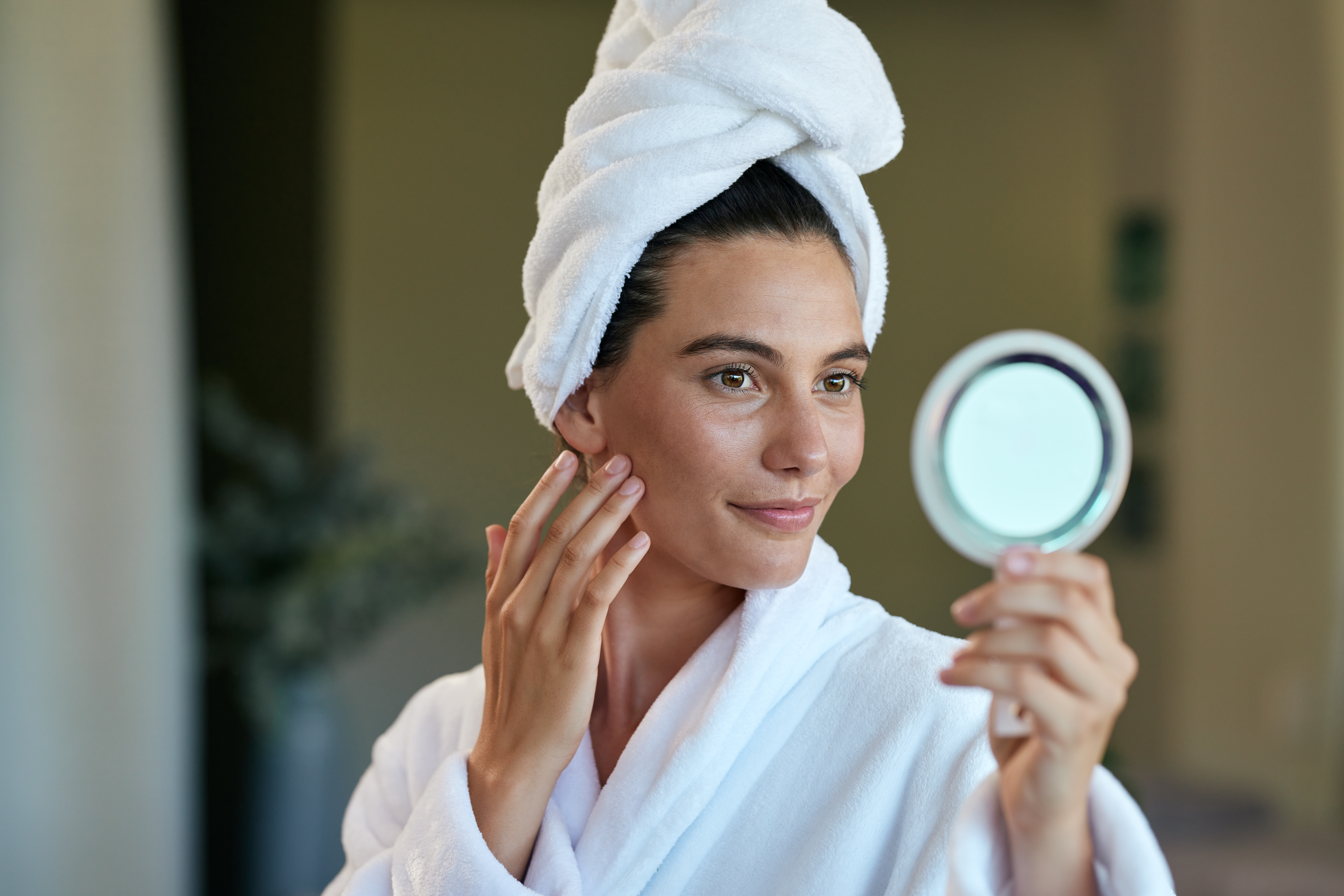 Mulher de pele morena, com toalha branca enrolada na cabeça e roupão branco, sorri enquanto toca o rosto e se olha em um espelho de mão redondo