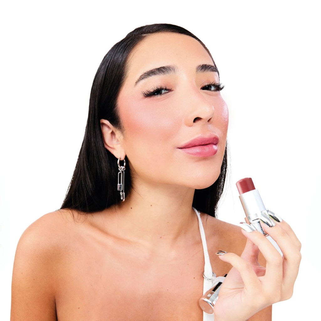 Mulher de cabelo preto liso, maquiagem rosa e brincos de alfinete, segurando um batom marrom avermelhado, sorrindo levemente para a c&acirc;mera