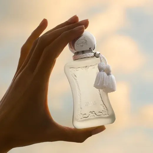 M&atilde;o feminina segura um frasco de perfume Parfums de Marly Valaya, com l&iacute;quido claro, tampa prateada e dois pingentes brancos, contra um c&eacute;u azul e laranja
