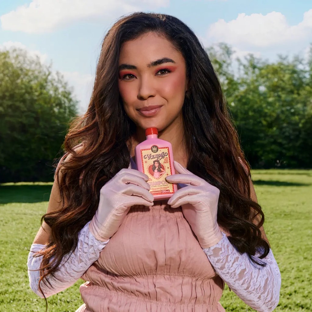 Mulher jovem com cabelo longo e escuro, maquiagem rosa nos olhos e bochechas, sorrindo levemente, segurando um frasco rosa de Volupto Shampoo com luvas de cetim e renda, em um campo verde sob c&eacute;u azul