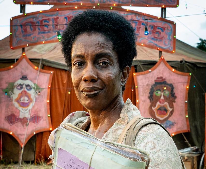 Mulher negra de cabelo crespo curto, vestindo blusa estampada clara, segura uma pilha de papéis amarrados com barbante, sorrindo levemente. Ao fundo, tenda de circo com letreiro iluminado e duas placas cor-de-rosa com rostos de palhaços desenhados