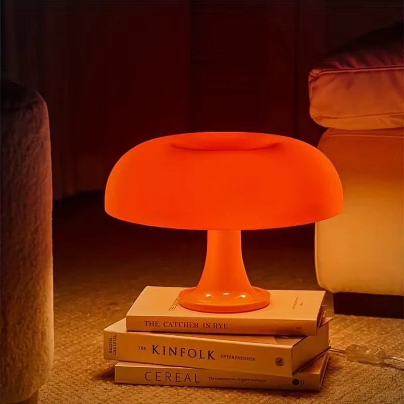 Sala de estar aconchegante com sof&aacute; bege, almofada e manta marrons. Uma lumin&aacute;ria de ch&atilde;o com c&uacute;pula clara ilumina o ambiente. &Agrave; esquerda, uma mesa de centro redonda com caneca, livro e vela acesa. Ao fundo, prateleira com objetos decorativos e planta em vaso.
