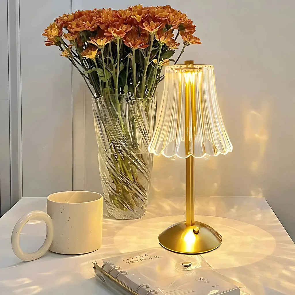 Mesa de cabeceira branca com um vaso de vidro transparente cheio de flores alaranjadas, uma lumin&aacute;ria dourada acesa com c&uacute;pula de vidro canelado, uma caneca bege e um caderno transparente com an&eacute;is dourados
