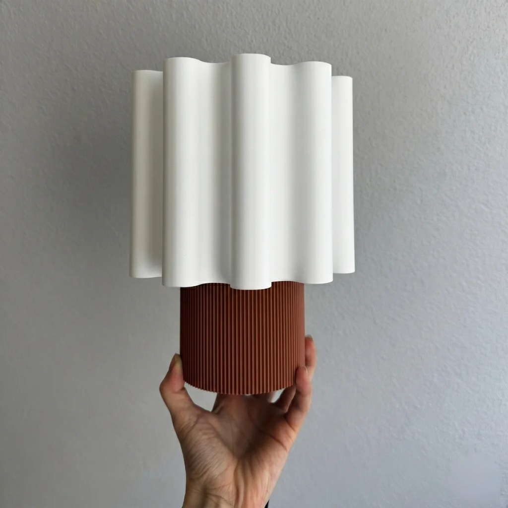 M&atilde;o feminina segura uma lumin&aacute;ria de mesa com base cil&iacute;ndrica marrom texturizada e c&uacute;pula branca ondulada, contra parede cinza clara