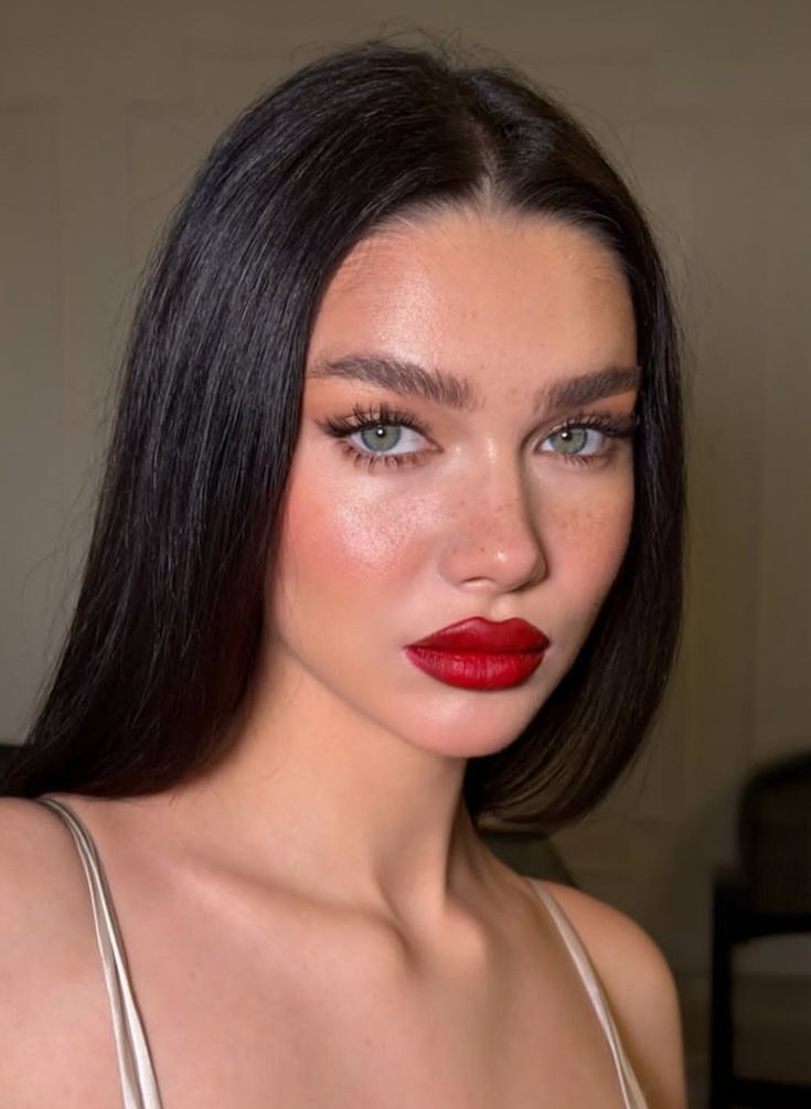 Mulher jovem com cabelos escuros e lisos, maquiagem marcante com batom vermelho vibrante, delineador e c&iacute;lios volumosos, e olhos verdes claros. Ela usa uma blusa de al&ccedil;as finas em tom claro e tem algumas sardas vis&iacute;veis no rosto