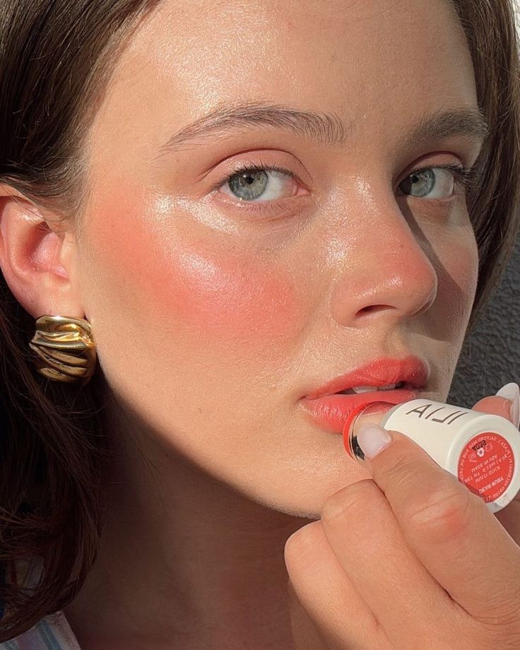 Mulher de pele clara, bochechas rosadas e olhos azuis aplicando batom coral nos l&aacute;bios, usando um brinco dourado ondulado