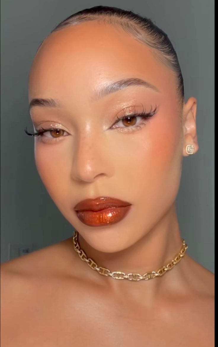 Rosto de uma mulher com maquiagem vibrante, pele bronzeada, l&aacute;bios com gloss marrom-avermelhado, olhos com sombra brilhante e delineado, bochechas rosadas, sobrancelhas definidas e um colar dourado de elos grossos