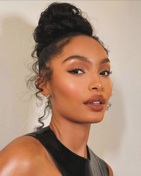 Mulher jovem com pele morena, cabelo escuro preso em coque alto, maquiagem natural com delineador gatinho e batom marrom, usando blusa preta de couro, olhando para frente com express&atilde;o serena