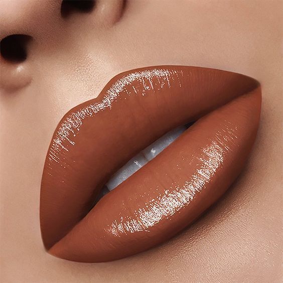 Boca de uma pessoa com l&aacute;bios volumosos e brilhantes, maquiados com batom marrom-claro e gloss, dentes brancos levemente vis&iacute;veis