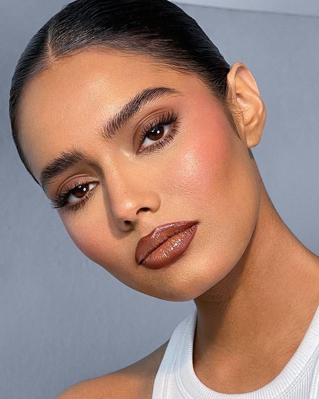Close-up de uma mulher com pele morena, cabelo escuro puxado para tr&aacute;s, maquiagem com blush rosado, l&aacute;bios com gloss marrom e c&iacute;lios longos. Ela veste uma blusa branca canelada