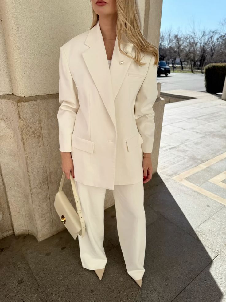Mulher loira, de p&eacute;, usando um elegante conjunto de blazer e cal&ccedil;a brancos, com uma bolsa creme e sapatos de bico fino da mesma cor, em um ambiente externo com luz solar