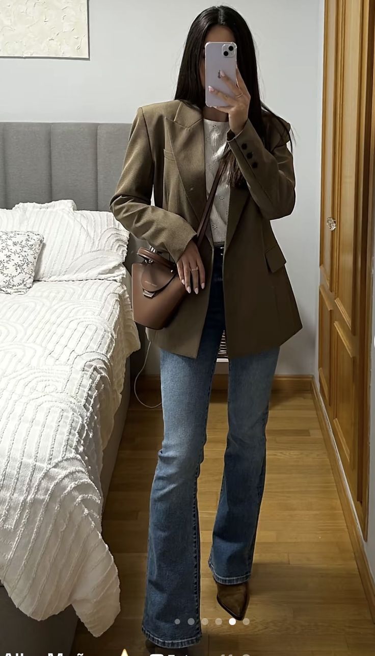 Mulher de cabelos longos e escuros tira selfie no espelho, vestindo blazer marrom, blusa branca, cal&ccedil;a jeans boca de sino e bota marrom. Ela segura uma bolsa pequena marrom e um celular lil&aacute;s, com um anel dourado no dedo. Ao fundo, uma cama com colcha branca texturizada e um guarda-roupa de madeira.